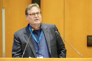 A man with glasses speaks at a podium, engaging an audience. He wears a dark suit and a blue shirt, with a microphone in front of him. The background features wooden panels, emphasizing a formal setting. The focus is on his expression and delivery.