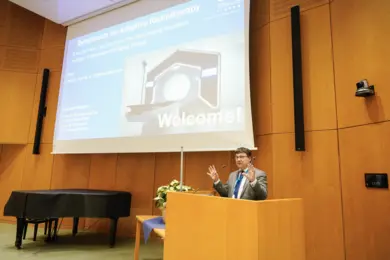 A speaker stands at a podium in a conference room, welcoming attendees to a symposium on Adaptive Radiotherapy. A projection screen displays the title and participant names, highlighting the event's focus on advancements in radiation treatment. The atmosphere is formal and academic.