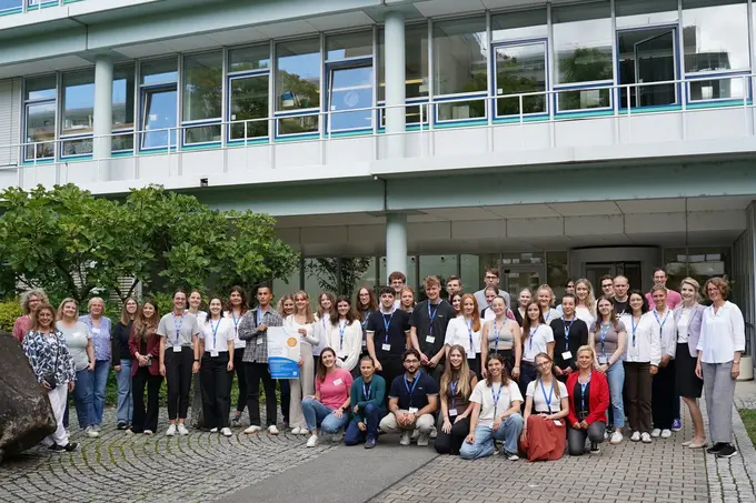 Eine große Gruppe von Menschen steht vor einem modernen Gebäude. Die Teilnehmer sind in unterschiedlichen Kleidungsstilen gekleidet und versammeln sich lächelnd für ein Gruppenfoto. Im Hintergrund sind Bäume und Fahrräder sichtbar, die zur Umgebung beitragen.