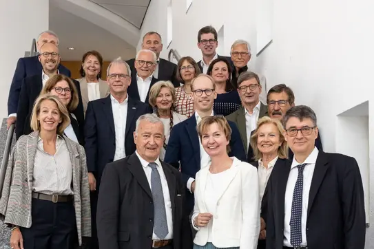 Gruppe von 17 Personen in formeller Kleidung steht auf einer Treppe und blickt in die Kamera. Die Personen tragen Anzüge, Blazer und elegante Kleidung. Der Hintergrund zeigt eine moderne Innenarchitektur mit weißen Wänden und kleinen Fenstern.