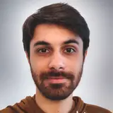 A young man with dark hair and a beard is smiling directly at the camera. He is wearing a two-toned hoodie, with the light gray part on top and brown on the bottom. The background is subtle and blurred.