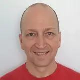 A smiling man with a shaved head stands against a plain background, wearing a red short-sleeve shirt. His hands are clasped behind his back, and he has a friendly expression.