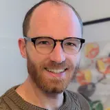 A smiling man with a short beard and glasses, wearing a knitted sweater, is facing the camera. He appears friendly and relaxed, set against a background with abstract artwork.