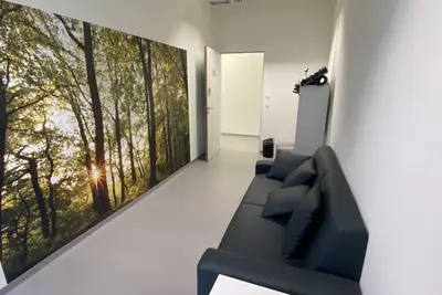Der Raum zeigt eine helle, moderne Umgebung mit einer großen Wandgrafik von Bäumen und Sonnenstrahlen. Auf der rechten Seite steht ein schwarzes Sofa mit Kissen, gegenüber befindet sich ein kleiner Tisch mit einigen Unterlagen. Eine offene Tür führt in einen angrenzenden Bereich.