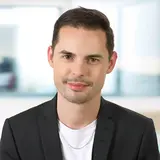 A young man with short dark hair and light skin, wearing a black blazer over a white shirt, smiles confidently. The background is a softly blurred interior space, creating a professional yet approachable atmosphere.