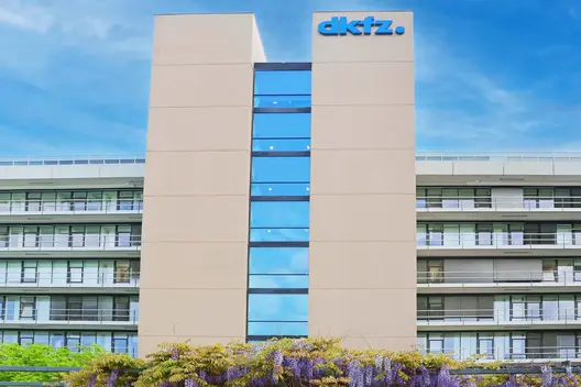 A modern, multi-story building features large glass windows and a prominent sign displaying the name "dkfz." The structure is surrounded by greenery and flowering plants, emphasizing an inviting environment. The sky is clear and blue, enhancing the overall appearance of the facility.