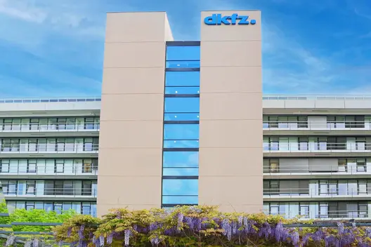 A modern, multi-story building features large glass windows and a prominent sign displaying the name "dkfz." The structure is surrounded by greenery and flowering plants, emphasizing an inviting environment. The sky is clear and blue, enhancing the overall appearance of the facility.
